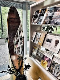 A modern interior featuring a bookshelf filled with various books and magazines, a large oval mirror reflecting part of the shelf and brick wall, and a potted plant in the foreground. The background shows a view of the exterior greenery through large windows.