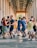 A group of people engaged in dancing under an arched walkway with columns on either side. The scene includes pairs of dancers moving energetically, with various expressions of joy and concentration. The background shows additional people casually observing the activity.