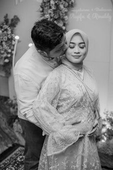 A couple is standing together in a loving embrace. The man is gently kissing the woman's cheek, while the woman, wearing a hijab and traditional clothing with intricate lace detailing, has her eyes closed and a peaceful expression on her face. The background is adorned with flower arrangements, enhancing the romantic atmosphere.