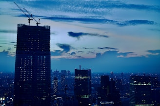 A panoramic shot of a modern high-rise building under construction at dusk, with cranes silhouetted against a darkening sky.