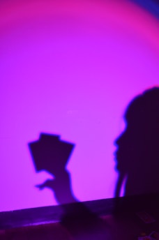 A shadow of a person holding two rectangular objects is visible against a brightly lit wall. The wall is bathed in a gradient of vibrant purple and pink hues, creating a dramatic contrast with the shadow.