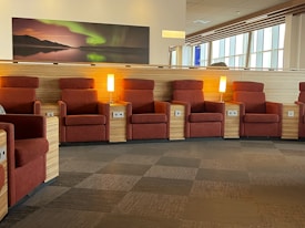 A series of comfortable, red lounge chairs are lined up alongside wooden partition walls. Small wooden tables with built-in power outlets and bright table lamps separate the chairs. Natural light streams in through large windows to the right, creating a warm and inviting atmosphere. A large photograph of a tranquil scene with the Northern Lights hangs above the chairs.