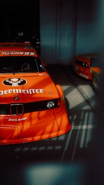 An orange race car, prominently featuring branding and logos, is mostly in the foreground with another car partially visible in the background. The setting appears to be a dimly lit garage or showroom, and there are noticeable shadows cast on the floor.