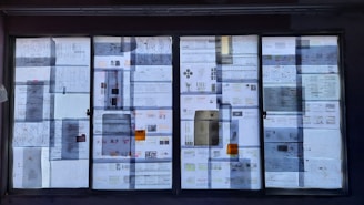 A large window is covered with various papers and documents, some with diagrams and charts. The papers are taped to the glass, creating an eclectic pattern with varying text and images, slightly blurred due to reflections and lighting.