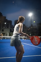 A person is playing padel on a court with a vibrant blue surface. They are holding an orange padel racket, preparing to hit a ball under bright overhead lights. The scene is set during dusk, with city buildings silhouetted in the background.