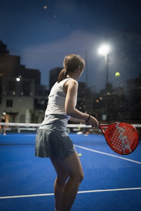 A person is playing padel on a court with a vibrant blue surface. They are holding an orange padel racket, preparing to hit a ball under bright overhead lights. The scene is set during dusk, with city buildings silhouetted in the background.
