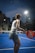 A person is playing padel on a court with a vibrant blue surface. They are holding an orange padel racket, preparing to hit a ball under bright overhead lights. The scene is set during dusk, with city buildings silhouetted in the background.