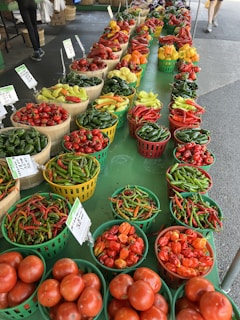 A vibrant market display featuring an array of colorful peppers and tomatoes. The vegetables are arranged neatly in baskets and containers on a green table. Signage indicates different prices and pepper types, and there are a few people browsing in the background.