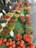 A vibrant market display featuring an array of colorful peppers and tomatoes. The vegetables are arranged neatly in baskets and containers on a green table. Signage indicates different prices and pepper types, and there are a few people browsing in the background.