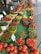 A vibrant market display featuring an array of colorful peppers and tomatoes. The vegetables are arranged neatly in baskets and containers on a green table. Signage indicates different prices and pepper types, and there are a few people browsing in the background.