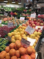 A bustling fruit and vegetable market showcases an array of colorful produce, including apples, persimmons, papayas, pears, and various leafy greens. Each type of produce is neatly arranged with handwritten signs displaying prices and names. The vibrant display is enhanced by overhead lighting, and the shop's signage and atmosphere convey freshness and variety.