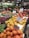 A bustling fruit and vegetable market showcases an array of colorful produce, including apples, persimmons, papayas, pears, and various leafy greens. Each type of produce is neatly arranged with handwritten signs displaying prices and names. The vibrant display is enhanced by overhead lighting, and the shop's signage and atmosphere convey freshness and variety.