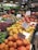 A bustling fruit and vegetable market showcases an array of colorful produce, including apples, persimmons, papayas, pears, and various leafy greens. Each type of produce is neatly arranged with handwritten signs displaying prices and names. The vibrant display is enhanced by overhead lighting, and the shop's signage and atmosphere convey freshness and variety.