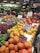 A bustling fruit and vegetable market showcases an array of colorful produce, including apples, persimmons, papayas, pears, and various leafy greens. Each type of produce is neatly arranged with handwritten signs displaying prices and names. The vibrant display is enhanced by overhead lighting, and the shop's signage and atmosphere convey freshness and variety.