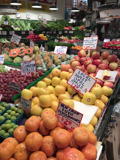 A bustling fruit and vegetable market showcases an array of colorful produce, including apples, persimmons, papayas, pears, and various leafy greens. Each type of produce is neatly arranged with handwritten signs displaying prices and names. The vibrant display is enhanced by overhead lighting, and the shop's signage and atmosphere convey freshness and variety.