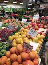 A bustling fruit and vegetable market showcases an array of colorful produce, including apples, persimmons, papayas, pears, and various leafy greens. Each type of produce is neatly arranged with handwritten signs displaying prices and names. The vibrant display is enhanced by overhead lighting, and the shop's signage and atmosphere convey freshness and variety.