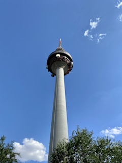 A tall telecommunications tower rises against a clear blue sky with a few white clouds. The base is surrounded by green trees, and the tower is capped with antennas and satellite dishes.