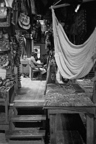 A cozy room filled with handcrafted textiles and woven items, including bags and hammocks. In the background, a person is seated at a table in a relaxed posture, surrounded by artisanal goods. The scene is dimly lit, highlighting the textures and patterns of the hanging cloth items.