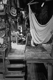 A cozy room filled with handcrafted textiles and woven items, including bags and hammocks. In the background, a person is seated at a table in a relaxed posture, surrounded by artisanal goods. The scene is dimly lit, highlighting the textures and patterns of the hanging cloth items.