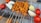 Skewers laden with colorful ingredients are lined up on a grill. Juicy chunks of marinated chicken, accompanied by whole red tomatoes and large pieces of white onion, are threaded onto metal sticks. The food is cooking over a layer of glowing charcoal, with smoke lightly wafting around.