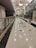 A long corridor with glossy marble flooring and geometric patterns, lined with glass walls on one side and a railing on the other. Overhead lights and chandeliers illuminate the space, providing a warm, inviting atmosphere. Large display windows are visible on the right side, possibly indicating a shopping area or commercial space.