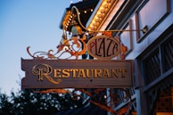 An ornate restaurant sign hangs outside in the evening, displaying the words 'Plaza Restaurant' with decorative elements and lights adorning the building facade.