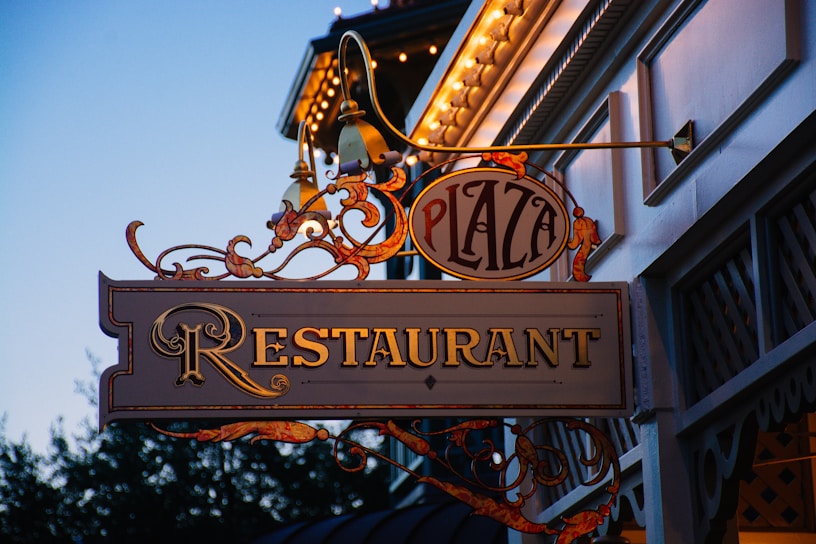 An ornate restaurant sign hangs outside in the evening, displaying the words 'Plaza Restaurant' with decorative elements and lights adorning the building facade.