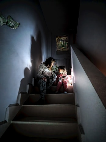 A woman and a child are sitting on a dimly lit staircase. The lighting creates a warm, intimate atmosphere. The woman appears to be engaged in conversation or storytelling with the child, who is clutching a stuffed toy. Shadows cast by the light enhance the cozy setting.