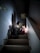 A woman and a child are sitting on a dimly lit staircase. The lighting creates a warm, intimate atmosphere. The woman appears to be engaged in conversation or storytelling with the child, who is clutching a stuffed toy. Shadows cast by the light enhance the cozy setting.