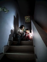 A woman and a child are sitting on a dimly lit staircase. The lighting creates a warm, intimate atmosphere. The woman appears to be engaged in conversation or storytelling with the child, who is clutching a stuffed toy. Shadows cast by the light enhance the cozy setting.