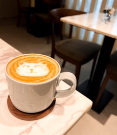 A mug of cappuccino is placed on a marble table with latte art depicting a cat's face. The table is set in a cozy cafe environment with wooden chairs and another table in the background.