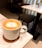 A mug of cappuccino is placed on a marble table with latte art depicting a cat's face. The table is set in a cozy cafe environment with wooden chairs and another table in the background.