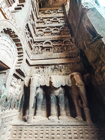 An intricate stone carving depicting an ancient architectural facade filled with arches, detailed sculptures, and two prominent elephant figures at the base. The craftsmanship showcases figures in various poses and elaborate decorative elements.