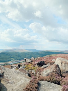 A vast landscape features rugged rocks and heather-covered hills under a partly cloudy sky. The rolling hills stretch into the distance with patches of forest visible below. A peaceful river flows through the valley, enhancing the scenic view.