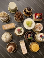 An assortment of intricately decorated pastries and cakes is displayed on a dark, textured surface. Each dessert is uniquely adorned with toppings like fruits, chocolate shavings, and powdered sugar, presented on gold-colored bases.