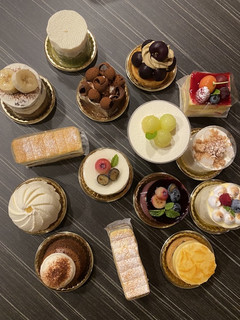 An assortment of intricately decorated pastries and cakes is displayed on a dark, textured surface. Each dessert is uniquely adorned with toppings like fruits, chocolate shavings, and powdered sugar, presented on gold-colored bases.