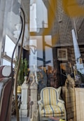 A store window display featuring a mix of vintage and eclectic furniture items. Prominent objects include a striped yellow and gray upholstered armchair, decorative pillars, an ornate mirror, and green plants. Reflections in the glass reveal city street signs and part of a building exterior.