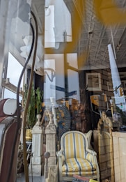 A store window display featuring a mix of vintage and eclectic furniture items. Prominent objects include a striped yellow and gray upholstered armchair, decorative pillars, an ornate mirror, and green plants. Reflections in the glass reveal city street signs and part of a building exterior.