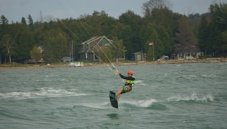 A kiteboarder wearing a helmet and a life vest is riding on choppy waters near a tree-lined shore. In the background, there are houses and a Canadian flag hanging from a pole.