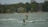 A kiteboarder wearing a helmet and a life vest is riding on choppy waters near a tree-lined shore. In the background, there are houses and a Canadian flag hanging from a pole.