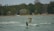 A kiteboarder wearing a helmet and a life vest is riding on choppy waters near a tree-lined shore. In the background, there are houses and a Canadian flag hanging from a pole.