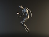 A stylized, metallic humanoid figure appears to be floating or suspended in mid-air against a dark background. The figure is interacting with a small, reflective sphere hovering near its hand.