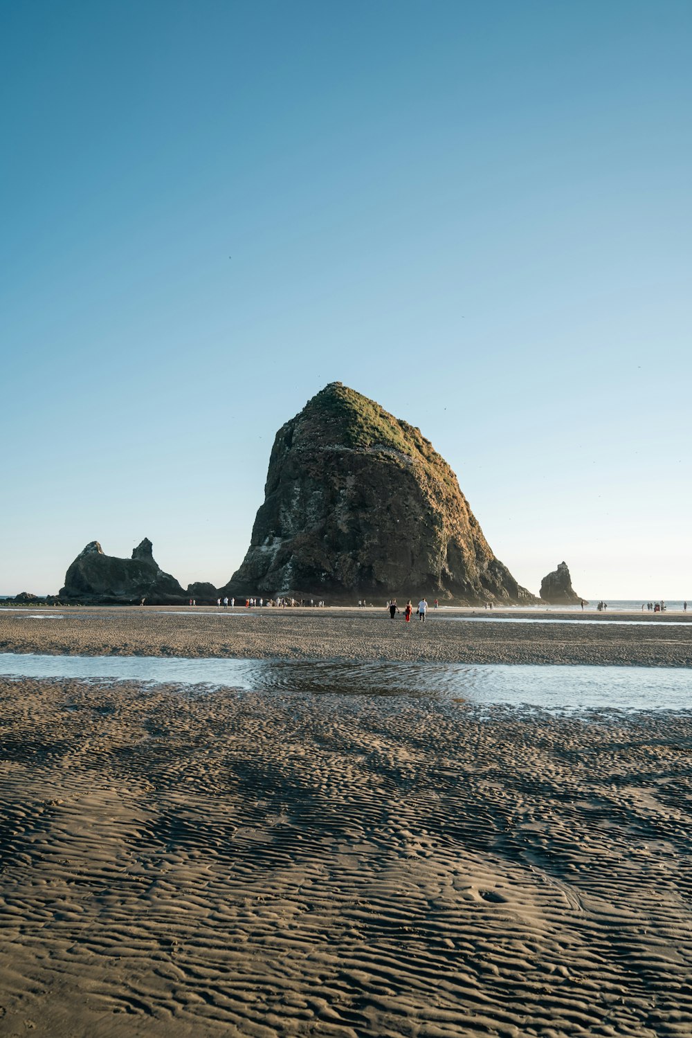 a group of people walking on a beach with large rocks in the background with Haystack Rock in the background