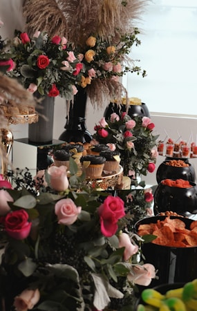 A lavish arrangement of pink, red, and yellow roses is set amid lush greenery and pampas grass in elegant vases. The setting features an array of snacks, including cupcakes on a decorative stand and bowls filled with various chips and snacks, creating a luxurious and inviting atmosphere.