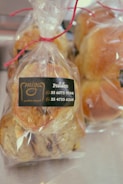 Transparent plastic bags containing freshly baked bread rolls are tied with red string. A label on the bag displays a logo and phone numbers for ordering.