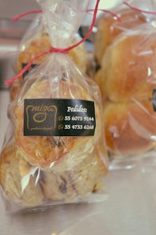 Transparent plastic bags containing freshly baked bread rolls are tied with red string. A label on the bag displays a logo and phone numbers for ordering.