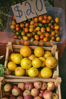 Crates filled with a variety of fruits are displayed, including red apples, yellow oranges, and small round fruits in shades of yellow, green, and orange. A handwritten sign with a price of $20 is placed on the crate of the small mixed-color fruits.