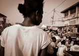 A person is seen from the back, wearing a white shirt, amidst a busy outdoor market setting. The scene captures a lively atmosphere with multiple people engaged in various activities. Stalls, products, and a variety of structures line the market, contributing to the bustling environment.