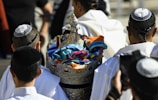 A group of men wearing traditional head coverings are gathered around a decorative silver container. The container is adorned with colorful fabrics and intricately detailed designs. The men are dressed in white and adorned with religious attire.