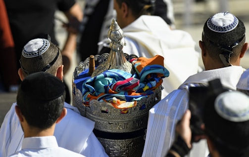 A group of men wearing traditional head coverings are gathered around a decorative silver container. The container is adorned with colorful fabrics and intricately detailed designs. The men are dressed in white and adorned with religious attire.